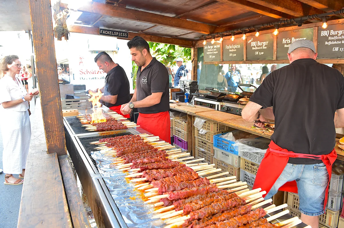 Fleischspiesse auf einem Grill. Eine Frau in weiss lässt sich von einem Verkäufer beraten.